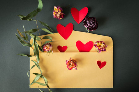 Romantic flat lay with yellow envelope, red paper hearts and dried roses on dark background....