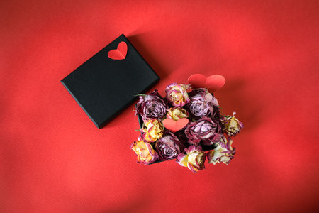 Black gift box with dried roses and red paper hearts on vibrant red background. Concept of faded...