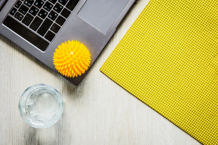 Home office wellness flat lay with laptop, green yoga mat, massage ball and water glass on wooden floor, calm remote work break and self care atmosphere