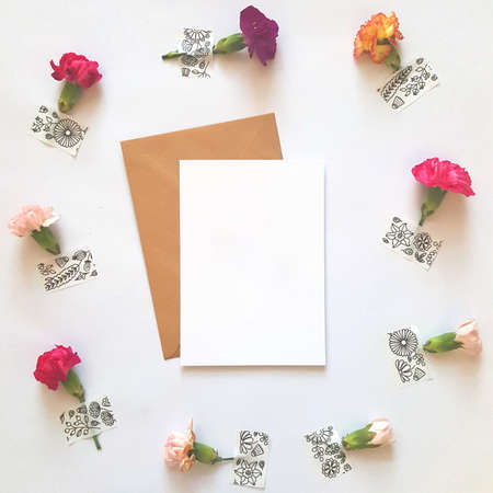 Blank card with brown envelope on white background with fresh flowers. Flatlay arrangement for...