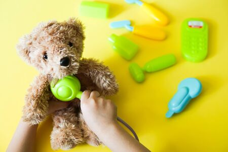 Child hands curing a bear. Kid toys medical equipment tool: stethoscope, thermometer, spoon, phone on yellow backgroundの写真素材