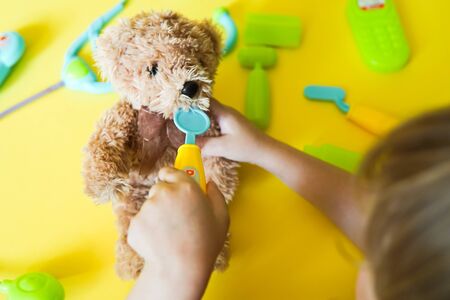 Child hands curing a bear. Kid toys medical equipment tool: stethoscope, thermometer, spoon, phone on yellow backgroundの写真素材