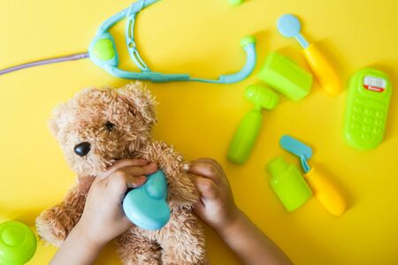 Child hands curing a bear. Kid toys medical equipment tool: stethoscope, thermometer, spoon, phone on yellow backgroundの写真素材