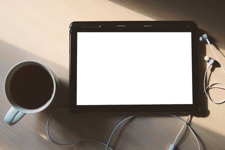 A blank tablet, earphones and a cup of tea on the wooden table. Pic in soft colors is perfect for...