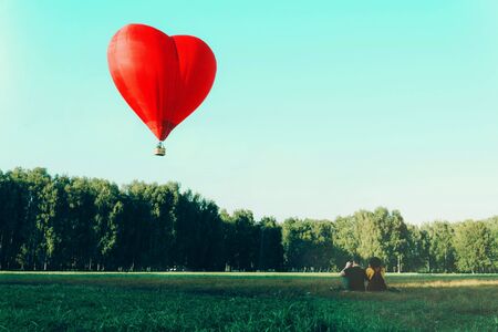 Young woman and man sitting on grass and looking at red hot air balloon in shape of heart. Love and future together concept.の写真素材