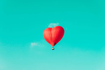 Red hot air balloon in the shape of a heart against the blue sky and some light clouds.の写真素材