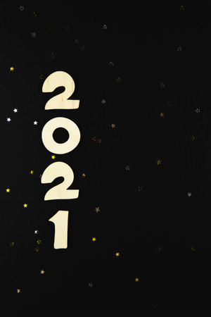 Wooden numbers 2021 on a black background with yellow and gray glitter stars. Copyspace, vertical.