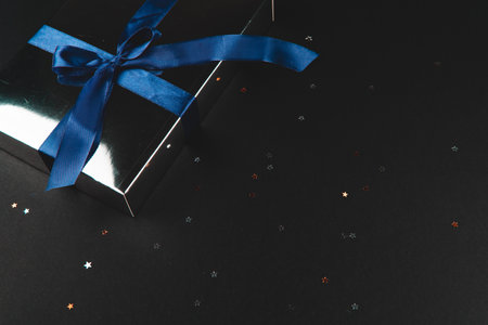 Gift box with a blue bow on a black background with small glitter stars. Holiday concept. Close up, flat lay, place for text.の写真素材
