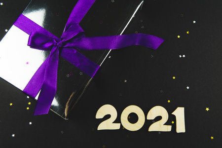 Gift box with a purple bow and numbers 2021 on a black background with small glitter stars. Holiday...