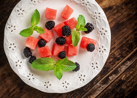 Refreshing summer salad with cubes of watermelon, blackberry, and mint on a white ceramic plate and a dark wooden background. Selective focus. Top viewの写真素材