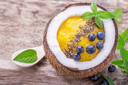Mango smoothies for breakfast with a garnish of chia seeds in the shell of a coconut on a wooden background with berries, blueberries and mint leaves. selective Focusの写真素材