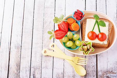 Lunch box for kids with fresh vegetables, fruits, nuts, berries and sandwich with cheese and herbs on a simple white wooden background. selective Focusの写真素材