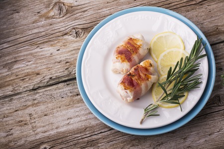 Chicken rolls with bacon grilled for a healthy diet with slices of fresh lemon and sprig of rosemary on a white plate on a simple wooden background with linen napkinの写真素材