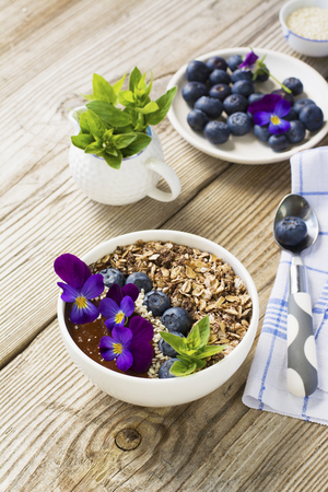 Chocolate Banana Smoothies Bowl with a topping of granola, blueberries, white sesame decorated with flowers purple garden viola on a simple wooden background. The concept of healthy organic breakfast snack.の写真素材