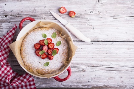 Home Sunday cake for dinner decorated with powdered sugar, strawberry slices and fresh mint leaves, basil. Served in a ceramic red background baking on a light wooden background with vintage tablecloth handmade.の写真素材