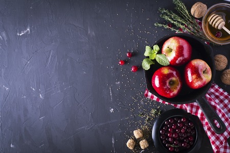 Juicy red fragrant apples for baking a dessert with cranberries, honey, nuts, mint, thyme and sugar cane and a black iron skillet on a dark background. Selective focus.の写真素材