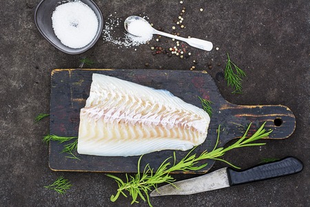 Raw cod before cooking on a black chopping Board with herbs and sea salt on a dark background. Top...