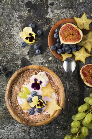 A healthy breakfast snack. Smoothies bowl with berry yogurt, fresh blueberries, carambola, figs, grapes, edible flowers in a wooden bowl on a gray stone background. Top Viewの写真素材