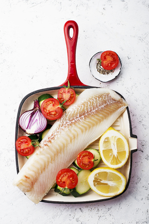 Cod. Fresh sea white raw fish before cooking in a frying pan with fresh vegetables: zucchini, sweet onions, tomatoes and lemons. Top view. Food concept.の写真素材