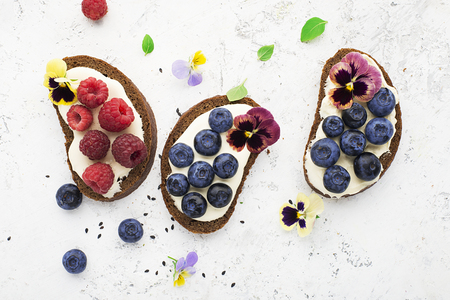 Light summer snack toasts with ricotta, honey, blueberries, raspberries and edible flowers of garden violets on dark-colored whole wheat bread on a light background. Top Viewの写真素材