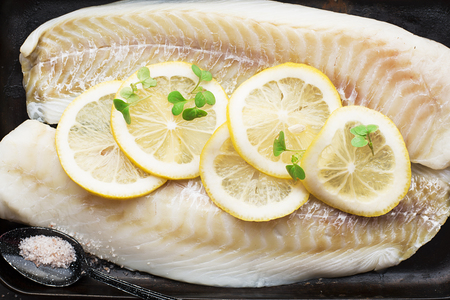 Raw cod fillets with lemon slices on a black baking tray. Top View