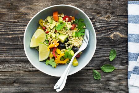 Healthy vegetarian lunch. Whole grain couscous, pastime pasta with fresh colorful fresh vegetables: bell peppers, cucumbers, avocados, olives, herbs, basil, olive oil. Top view.の写真素材