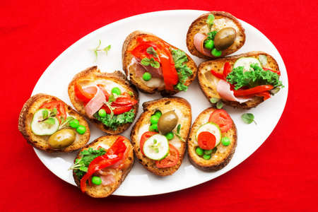 Assorted crostini canapes with cottage cheese, baked capsicum, cucumber slices, bacon, tomatoes, green peas on a festive red background. Top view,の写真素材