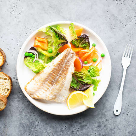 Fried fillet of sea white cod fish with juicy lettuce, capsicum, lemon, green peas on a large white dish on a gray background. Healthy balanced food. Top view,の写真素材
