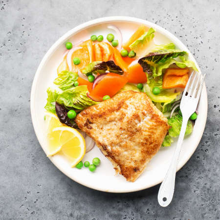Fried fillet of sea white cod fish with juicy lettuce, capsicum, lemon, green peas on a large white dish on a gray background. Healthy balanced food. Top view,の写真素材