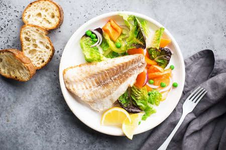 Fried fillet of sea white cod fish with juicy lettuce, capsicum, lemon, green peas on a large white dish on a gray background. Healthy balanced food. Top view,の写真素材