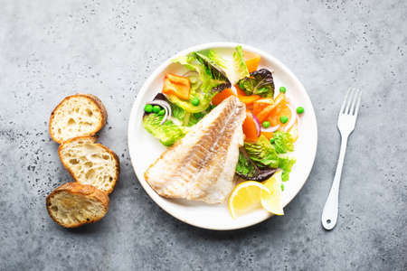 Fried fillet of sea white cod fish with juicy lettuce, capsicum, lemon, green peas on a large white dish on a gray background. Healthy balanced food. Top view,の写真素材