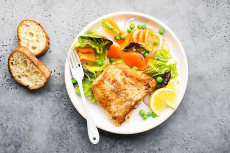 Fried fillet of sea white cod fish with juicy lettuce, capsicum, lemon, green peas on a large white dish on a gray background. Healthy balanced food. Top view,の写真素材