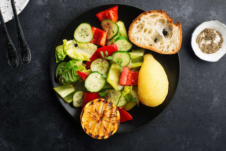 Young boiled potatoes, salad of leafy greens, cucumber, capsicum, micro greens, oil, with lemon on a dark background in a black plate with a slice of baguette. Top view.の写真素材