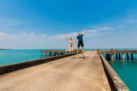 Young caucasian couple jumping on summer landscape with concrete Dai Mai pier on tropical koh Chang island  in Thailand. Seascape taken on honeymoon.の写真素材