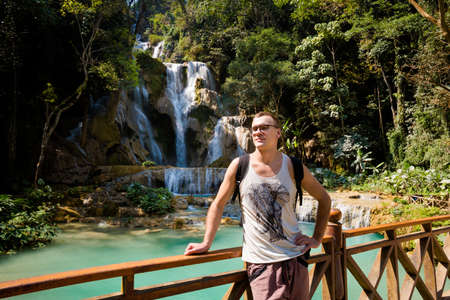 Young caucasian man on amazing Kuang Si waterfall near touristic Luang Prabang in Laos. Old wooden bridge and landscape of nature in south east asia during summer.の写真素材