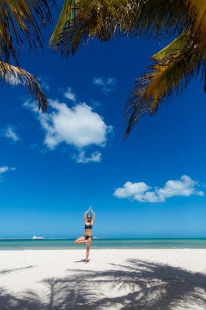 Beautiful young woman exercising yoga, tree pose, on a Progreso beach Mexico during sunny day. White beach with palms and blue sky.の写真素材