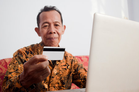 Old man hold and look at the credit card doing payment online with smart phone in living room, daily financial activityの写真素材