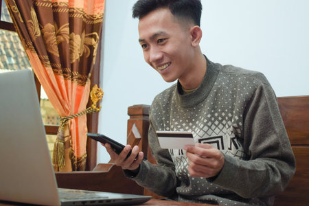 young man shopping online in e commerce, holding credit card and smartphone with laptop om table in livingroomの写真素材