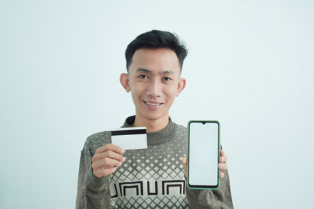 Joyful asian young men wear sweater holding blank white card and smartphone over white backgroundの写真素材