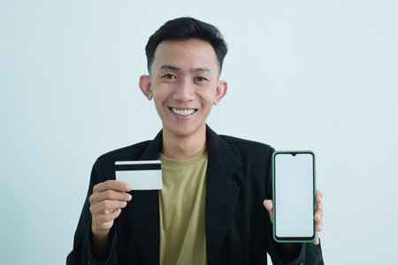 Joyful asian young businessman wear suit with tshirt holding blank white credit card and smartphone...