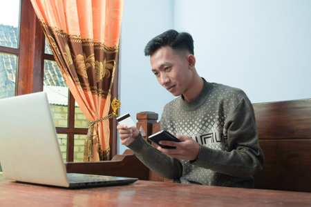 young man shopping online in e commerce, holding credit card and smartphone with laptop on table in living roomの写真素材