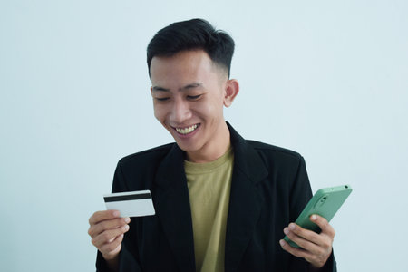 Joyful asian young businessman wear suit with tshirt holding blank white credit card and smartphone...