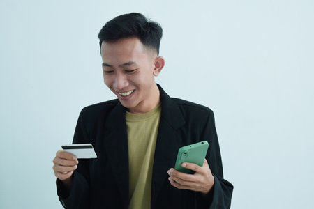 Joyful asian young businessman wear suit with tshirt holding blank white credit card and smartphone over white backgroundの写真素材