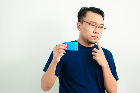Thoughtful asian man hold and pointing credit card. Isolated, blue color concept.の写真素材