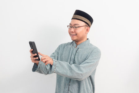 Asian Muslim man wearing clothes smiling holding phone pose, Ramadan Eid Al Fitr and eid adha days celebration isolated white background. Islamic celebration concept.の写真素材