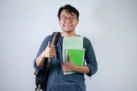 Asian young male student wearing bag and holding book smiling and happy expression. Ok hand sign gesture with isolated white backgroundの写真素材