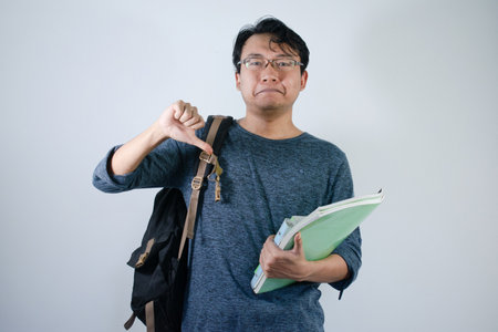 Asian young male reading book and making thumb down gesture. Male Student with glasses sad expression with Isolated white backgroundの写真素材