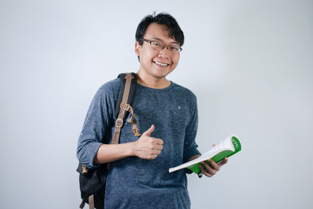 Asian young male student wearing bag and holding book smiling and happy expression. Ok hand sign gesture with isolated white backgroundの写真素材