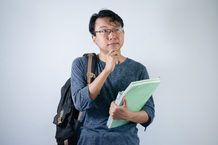 Asian young male reading book with thinking gesture. Asian male with glasses wearing grey shirt with...