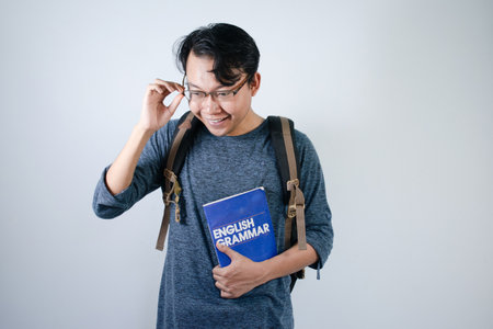 Asian young male student wearing bag and holding book smiling and hand on glasses. Isolated white backgroundの写真素材